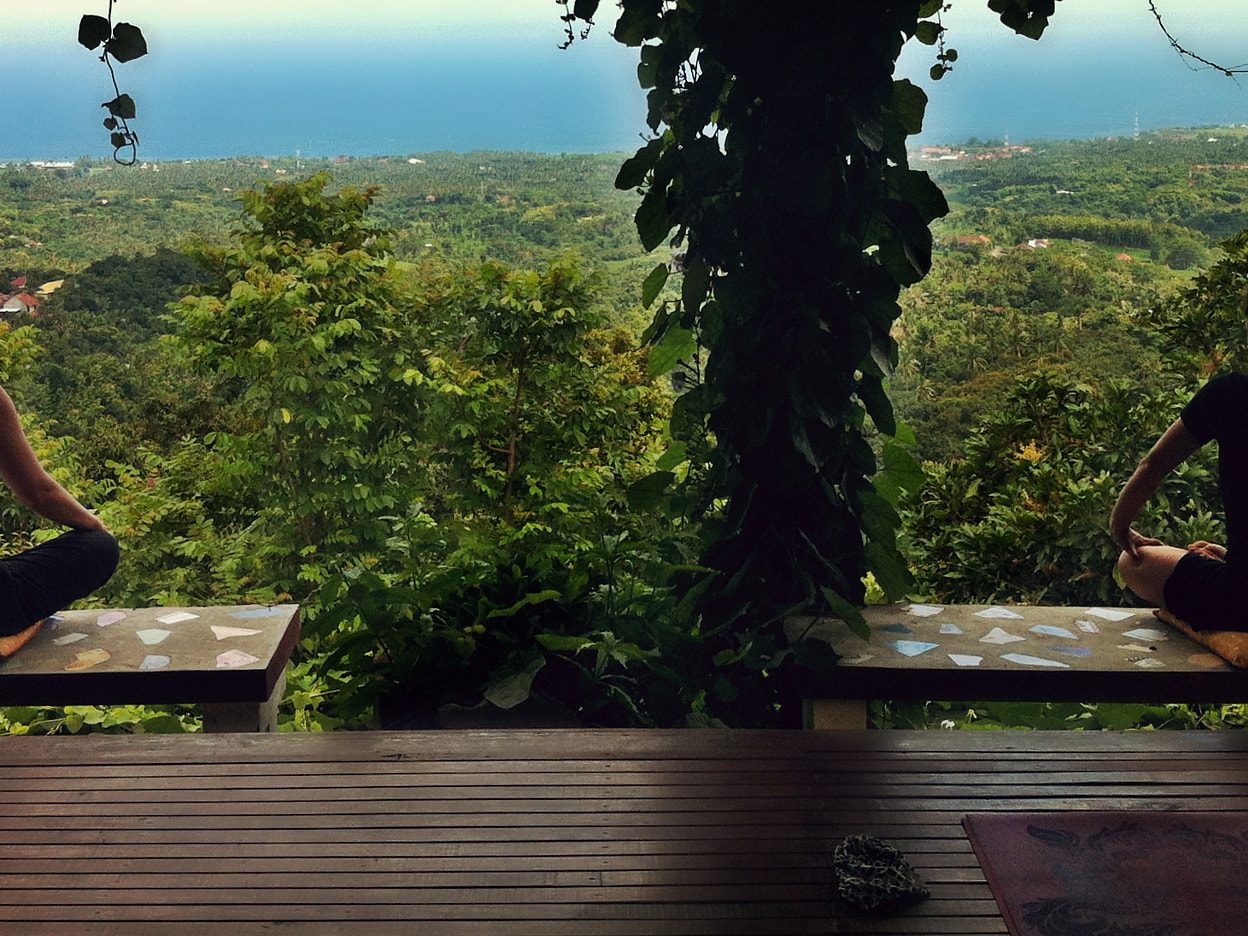 Zwei Menschen sitzen im Schneidersitz auf einer überdachten Holzterrasse, davor fällt der Blick über dichtes Grün bis zum Meer.